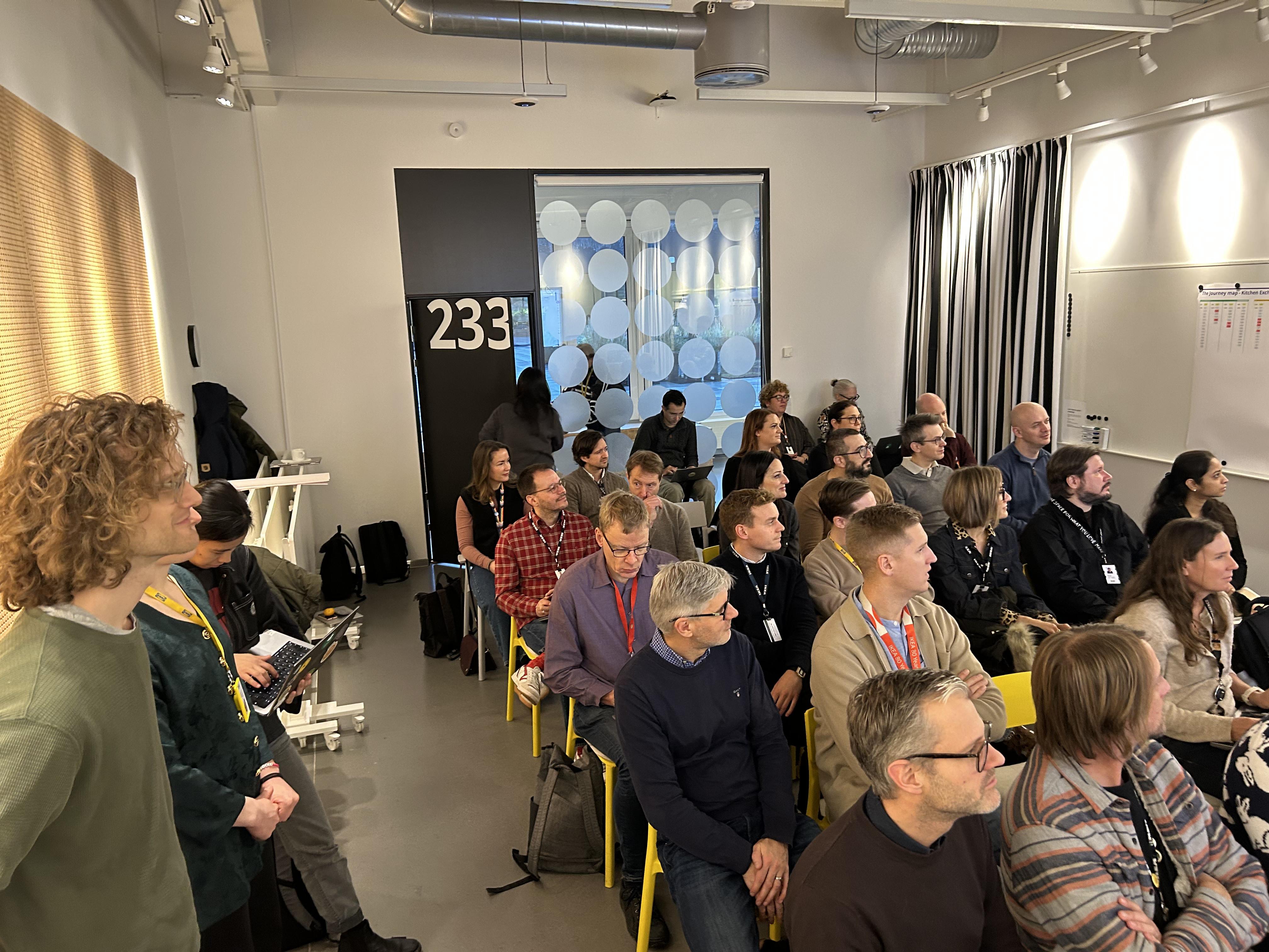 A packed workshop room of coworkers watching a presentation, with a printed journey map on the wall.